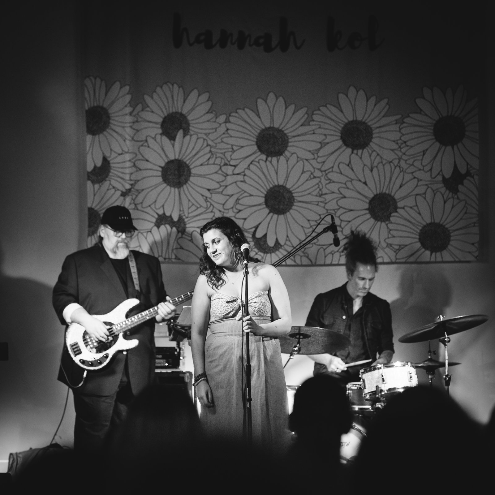 Vocalist on stage with bassist and drummer, a large painted-daisies backdrop behind her with her name in cursive script