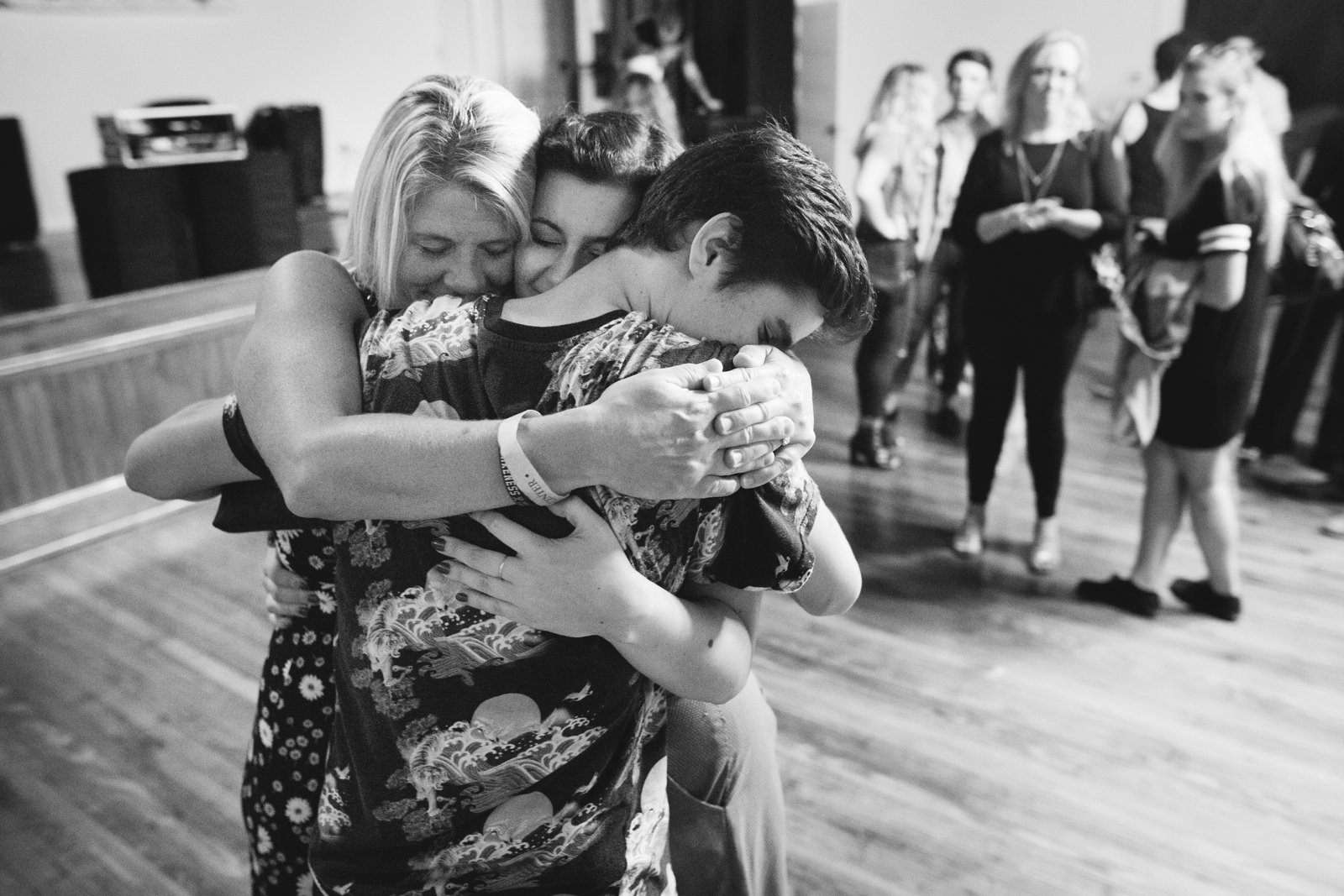 Black-and-white photograph of three people holding each other in a tight embrace on the venue floor after the show