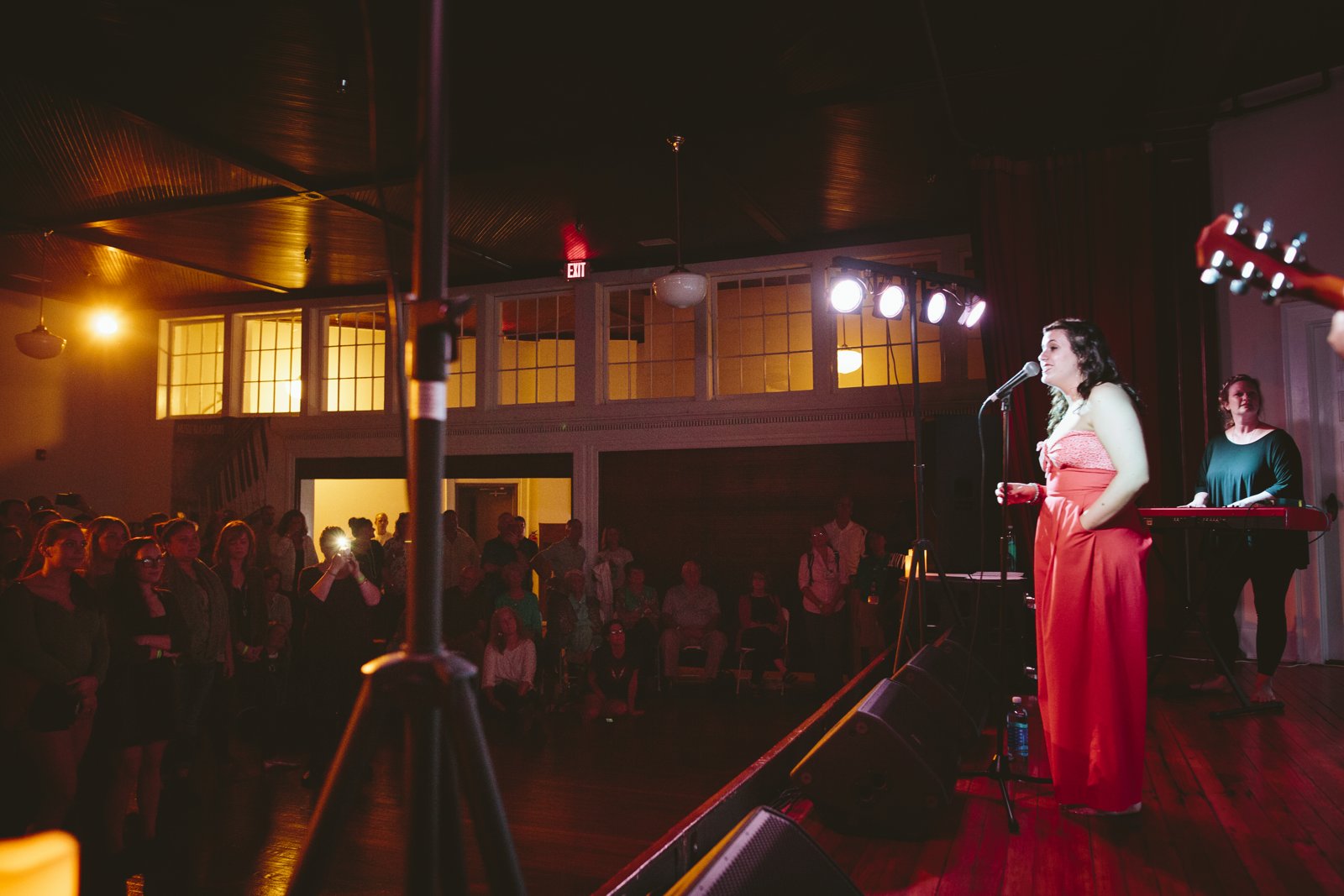 Vocalist in a red dress at the mic on a lit stage, keyboardist behind her, full audience gathered in the open venue